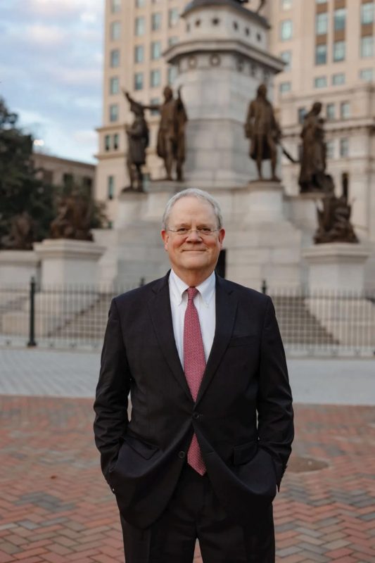 A man with glasses in a suit stands in front of a monument with bronze sculptures around it's base
